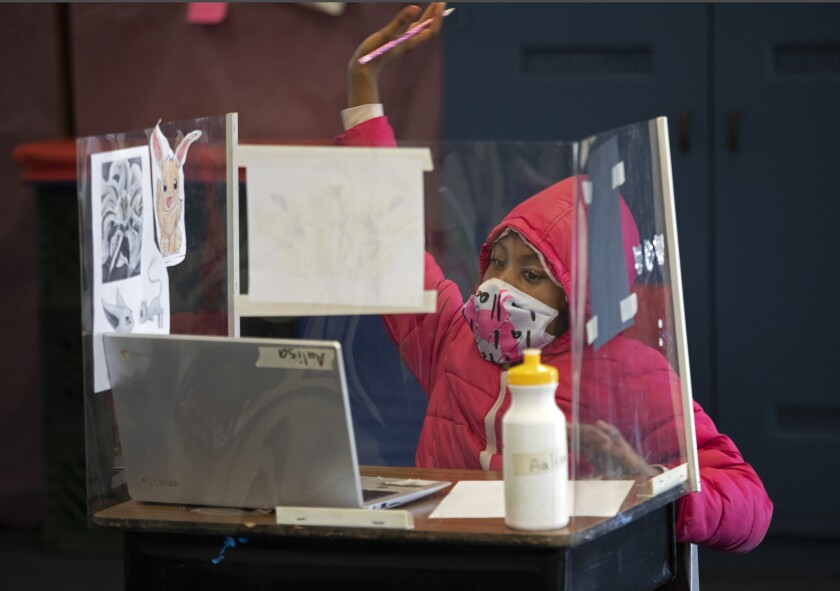 A child in a mask sits at a desk with a laptop and acrylic plastic dividers.