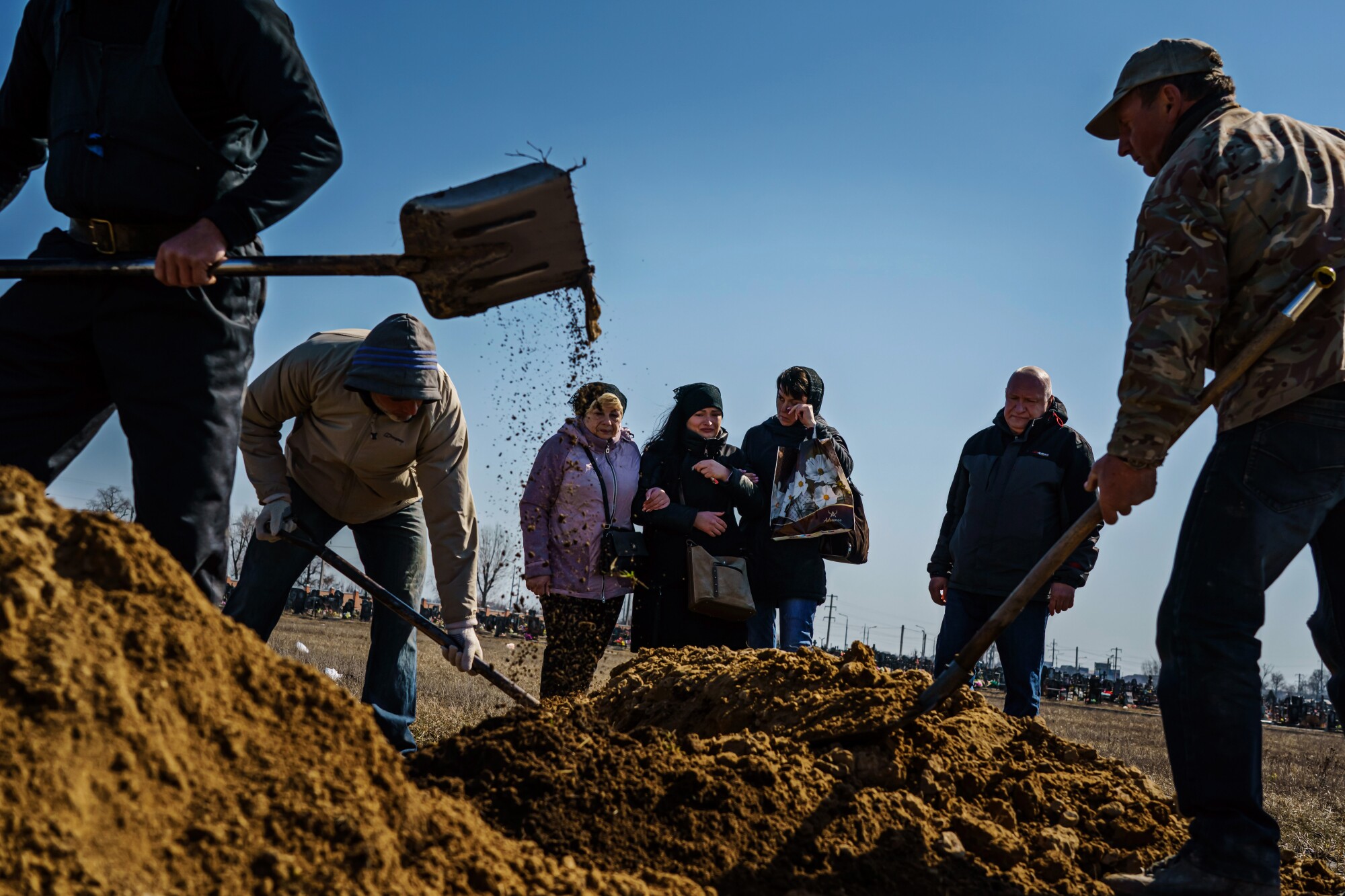 Three workers shoveling soil into a fresh grave as four mourners stand by.