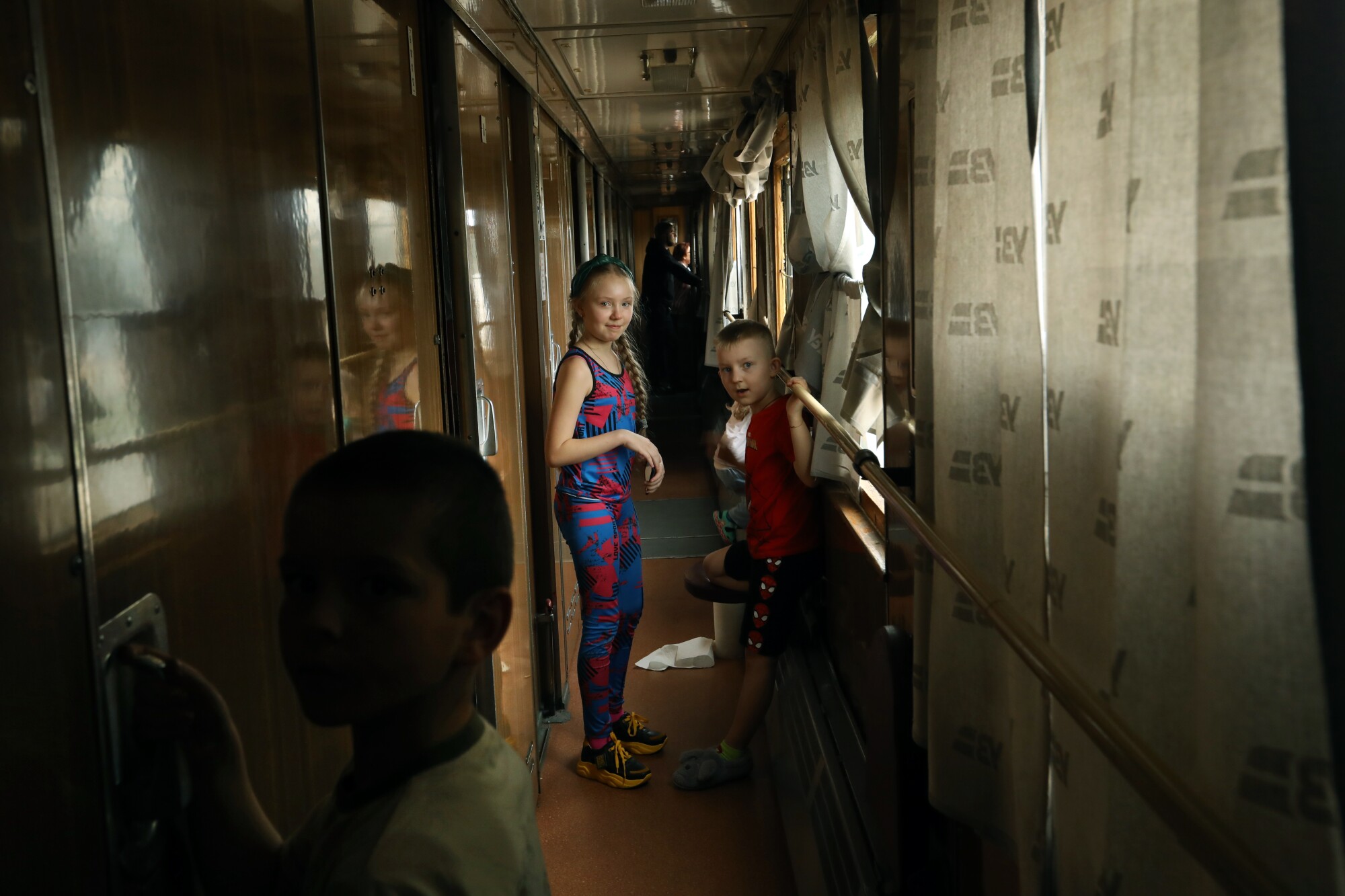 A girl and a boy stand in the train corridor while, in the foreground, a boy holds onto a door handle 