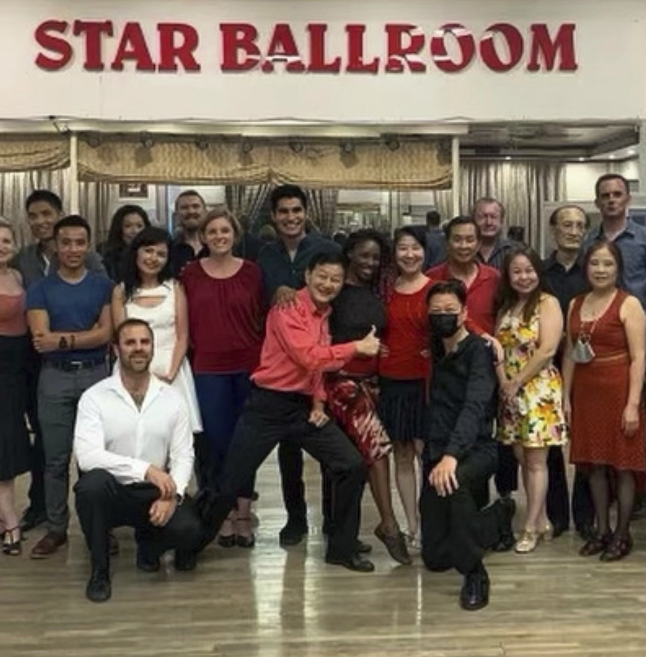 A photo of Mr. Ma, smiling and giving a thumbs up in a red shirt, with a group of community members at a dance event.