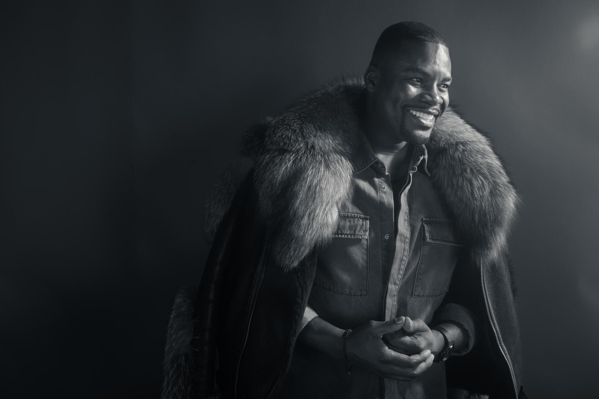 A man with a fur lines coat over his shoulders smiles.