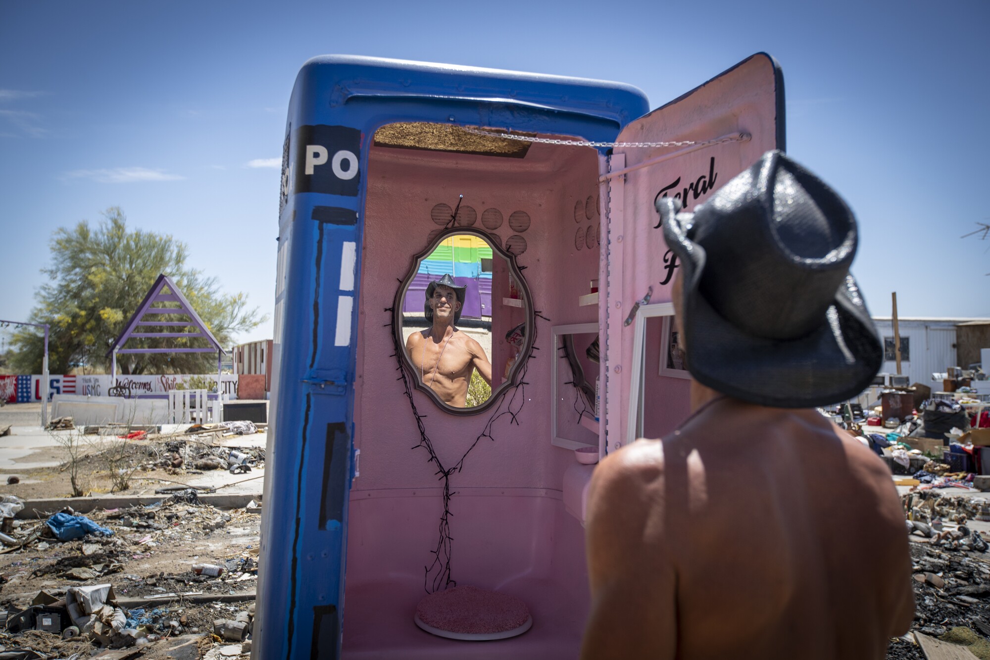 A shirtless man in a cowboy hat looks into a mirror inside a pink and blue painted portable toilet