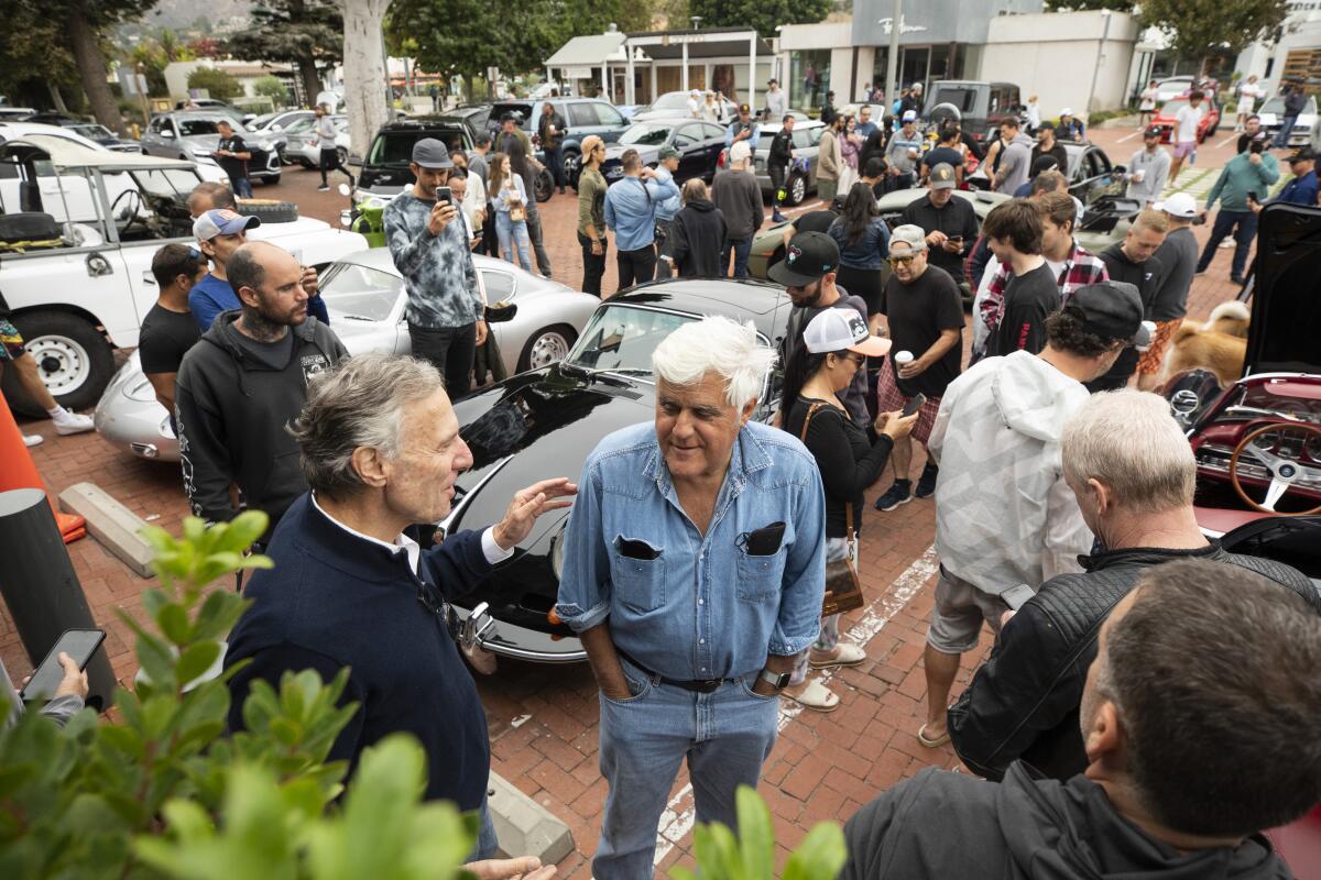 Jay Leno chats with a man in the foreground and lots of men mingle around cars in the background.