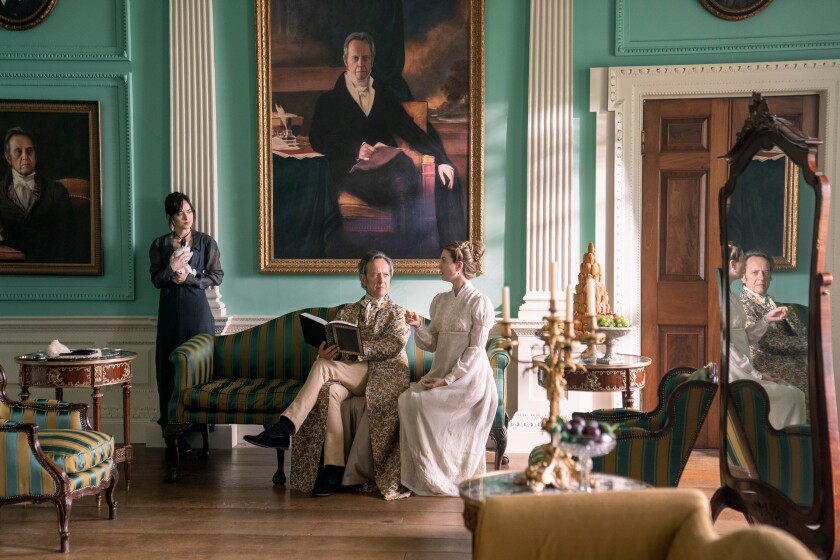 A man and woman are seated on a lavish living room couch. Another woman stands off to the left.