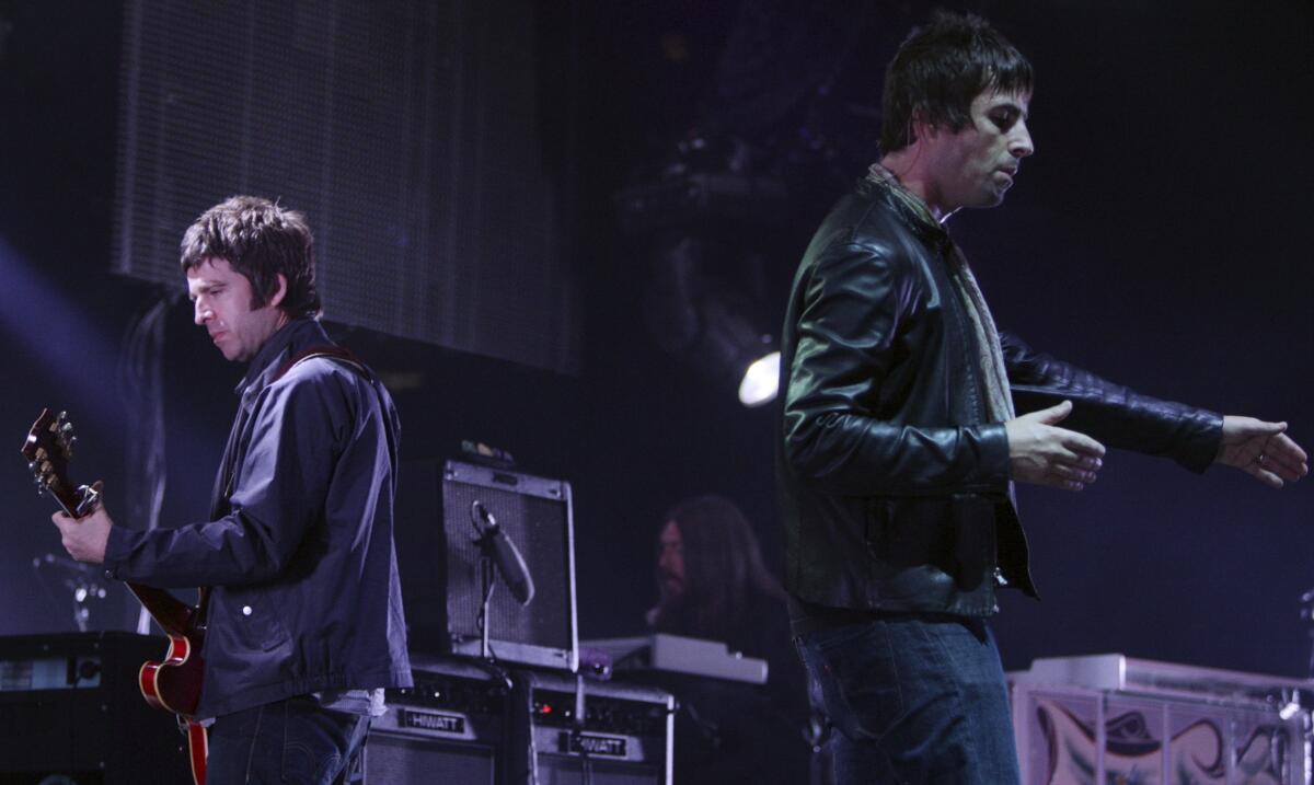 Noel Gallagher playing guitar while facing away from his brother Liam Gallagher onstage surrounded by equipment.