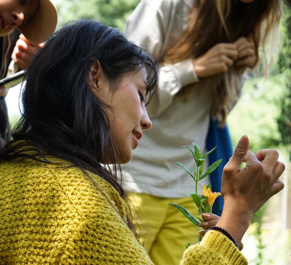 A person holds and points at a plant with a yellow bloom. Folks in the background observe.