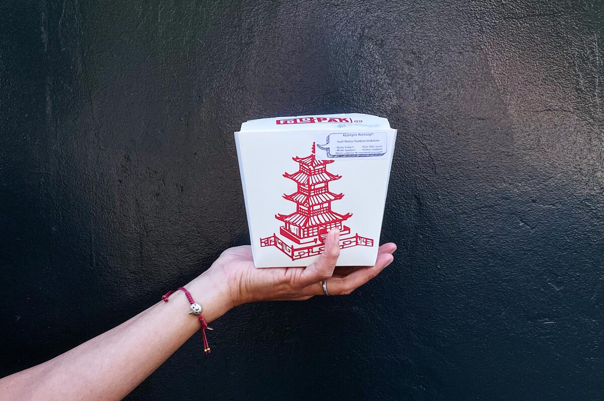 A hand holds a Chinese to-go container containing the frozen garlic noodles against a black wall.