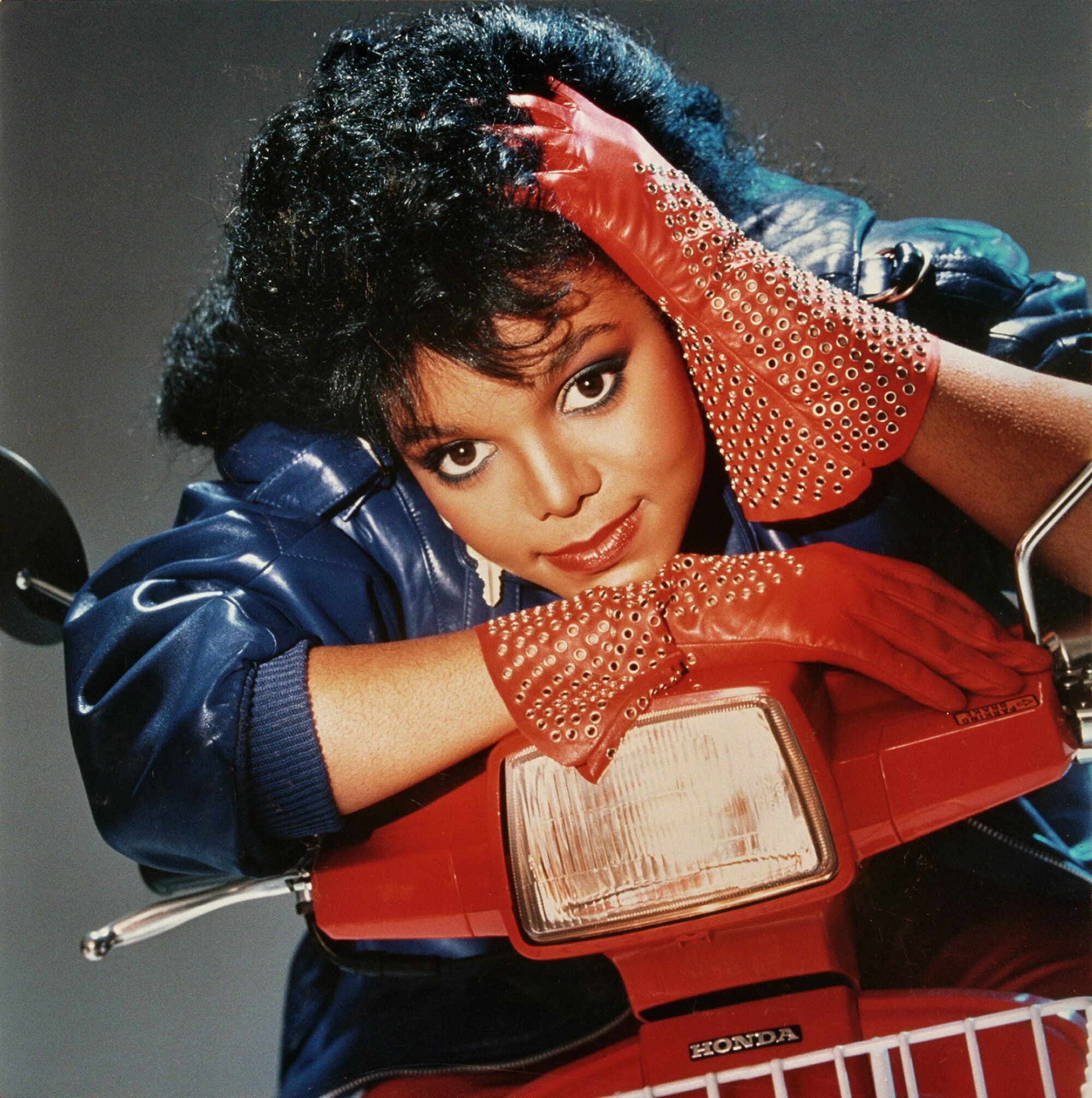 A young woman with red leather gloves rests her head on a red motorcycle