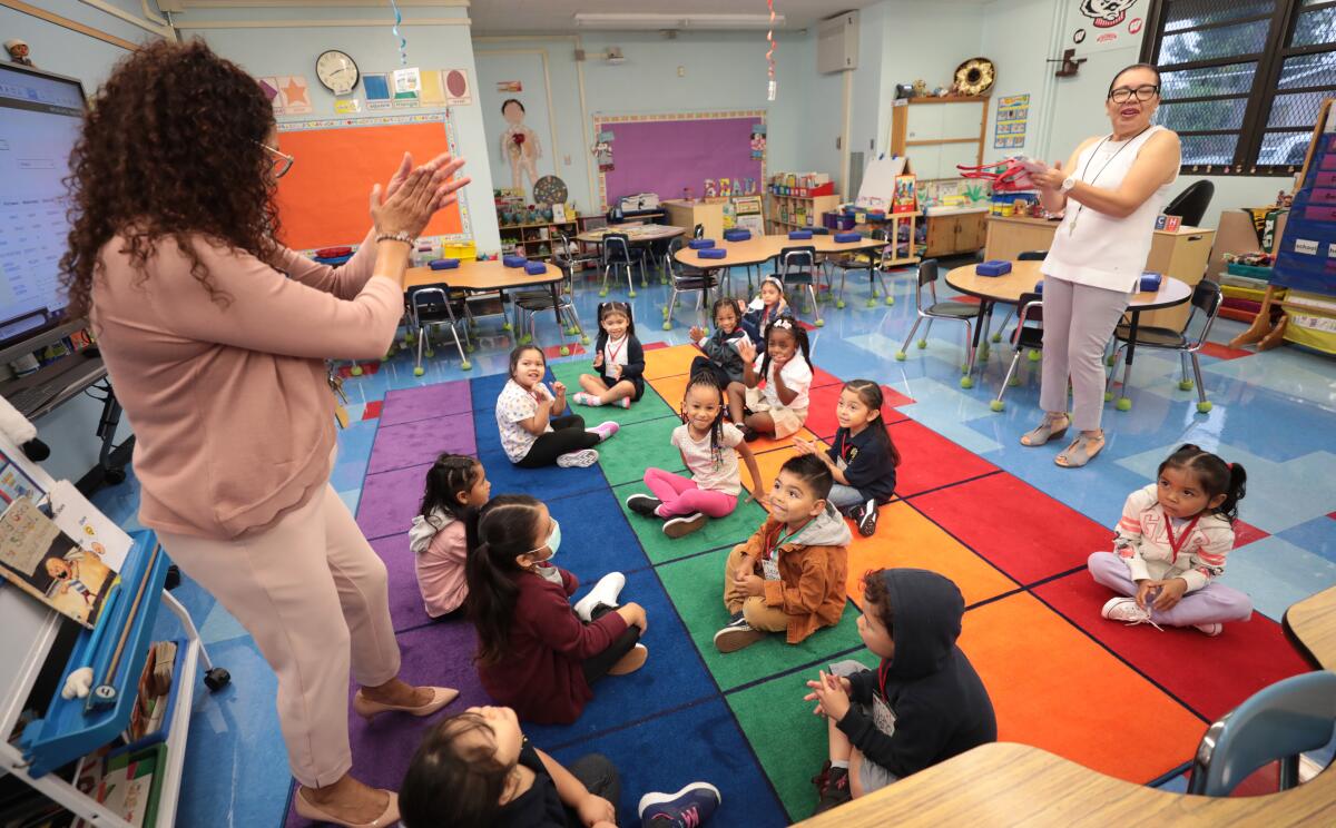 Transitional kindergarten students sit on a rainbow classroom rug and clap with their teacher and coach, who are standing.