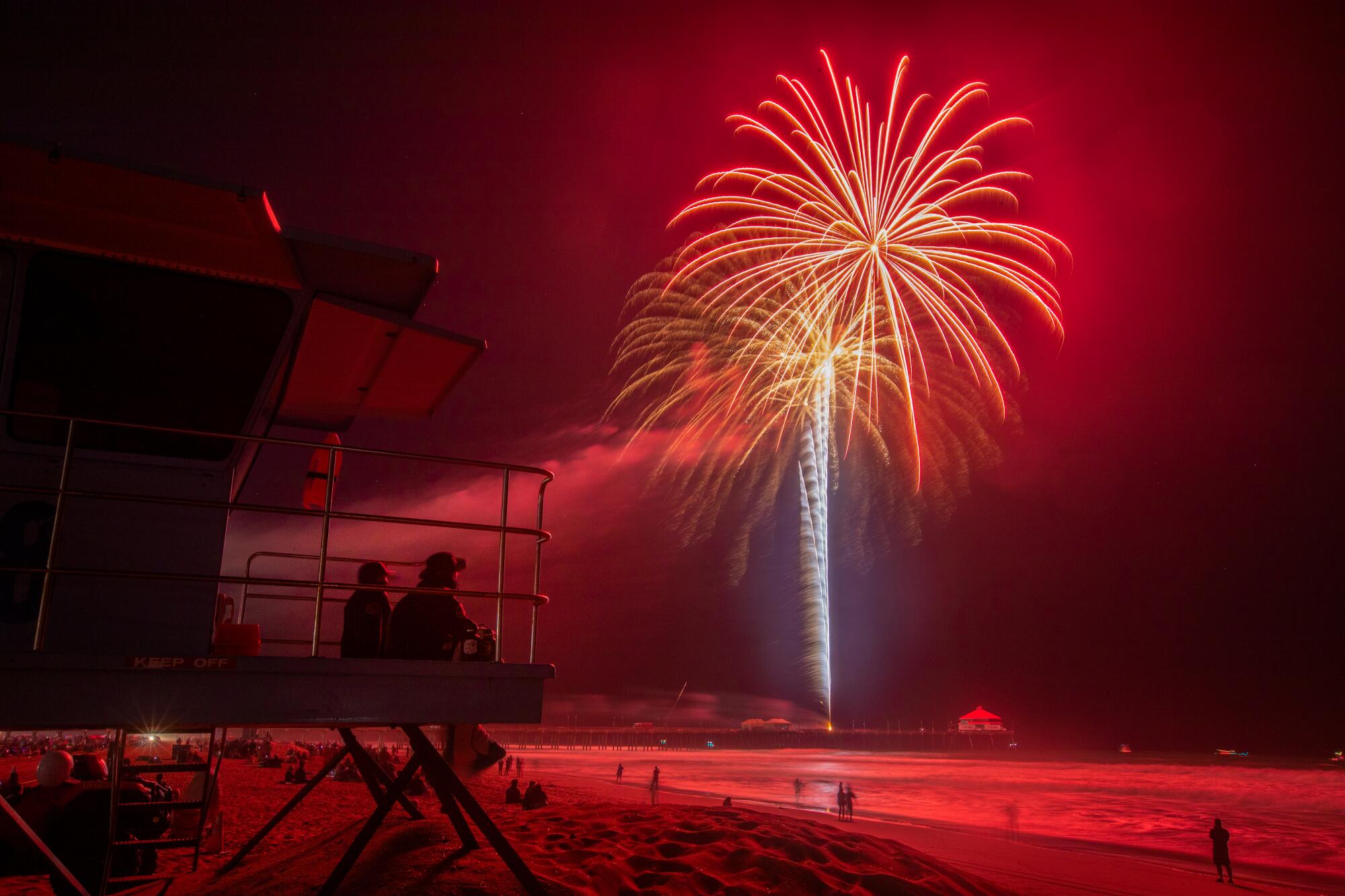 Two people sitting at a lifeguard tower watch a shower of pink and orange sparks from fireworks over the ocean