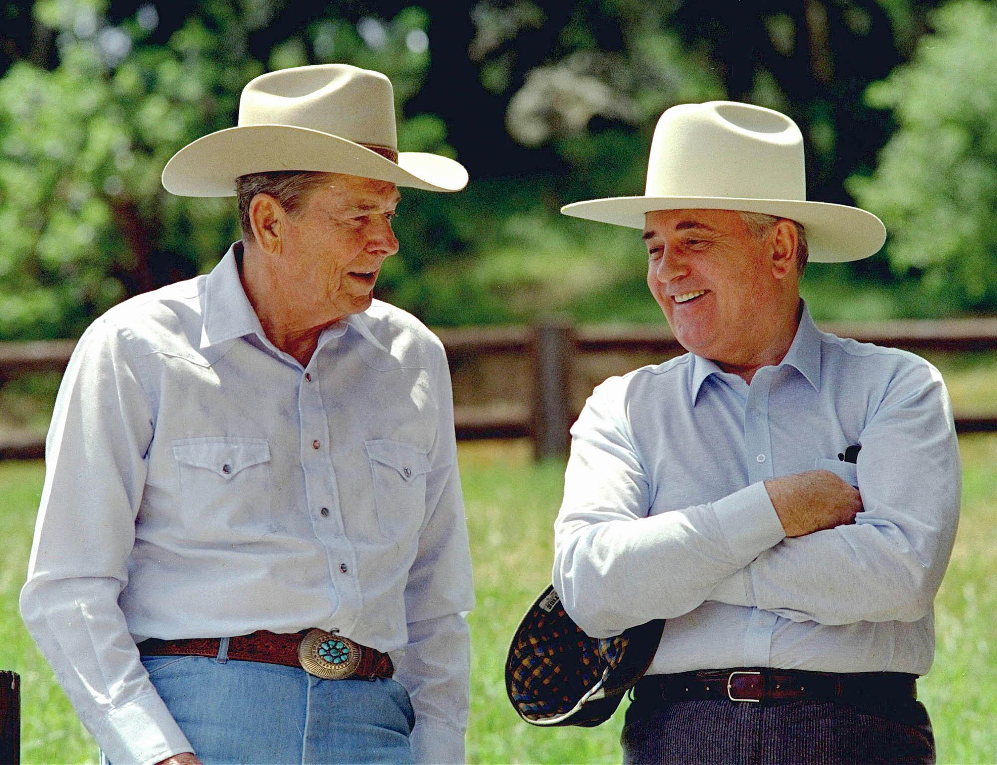 Two men in cowboy hats, one with his arms crossed and smiling on the right, share a conversation