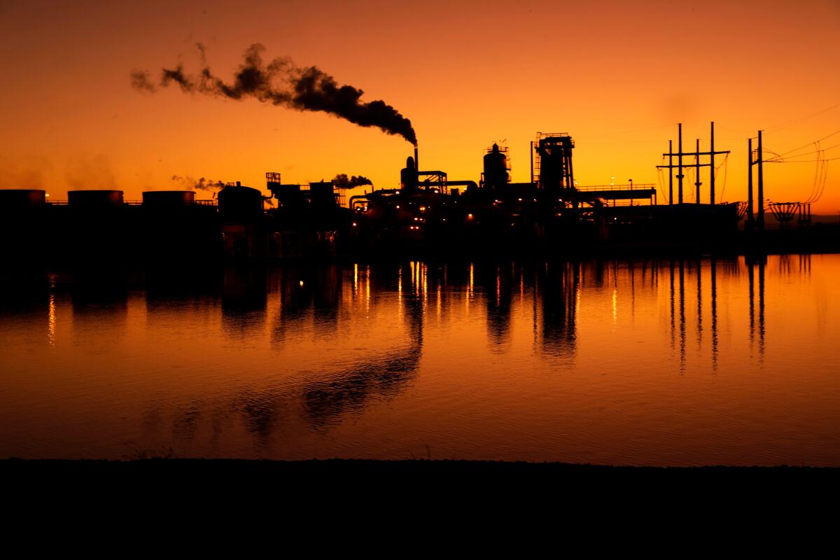 Smoke and industrial buildings are seen in silhouette at sunset and are reflected in a body of water.
