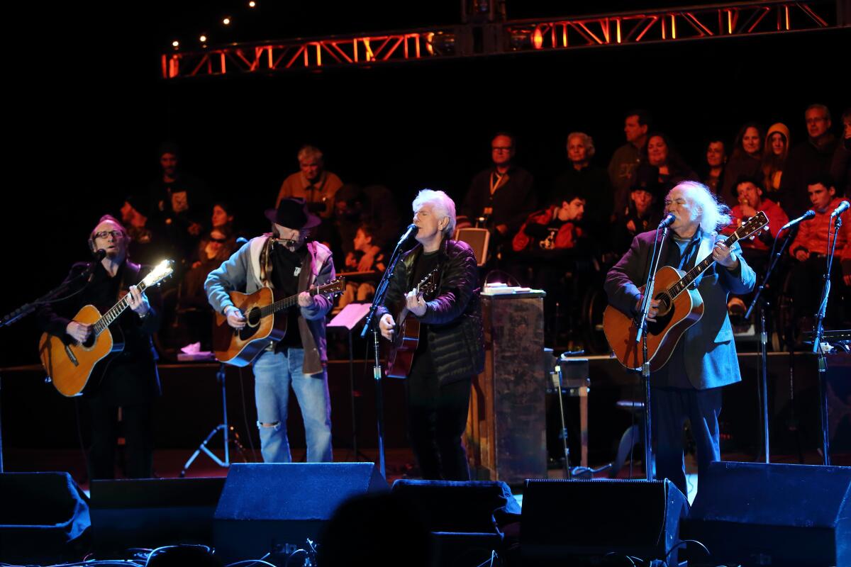 Crosby, Stills, Nash and Young playing guitars and singing into microphones on a stage.