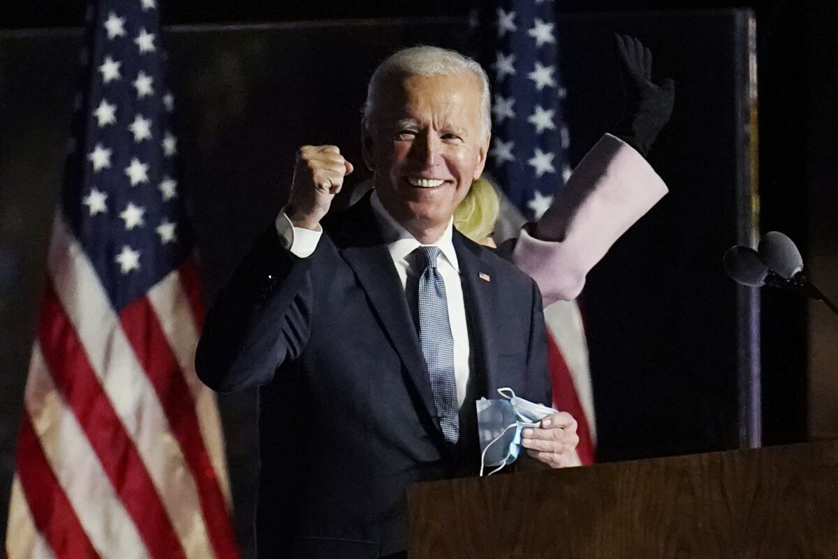 President Biden, mask in one hand, smiles and raises a fist with two U.S. flags in the background