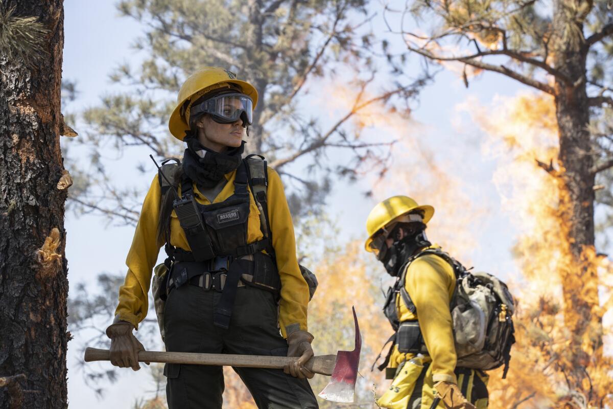 Angelina holds an ax by a tree while wearing firefighter gear.