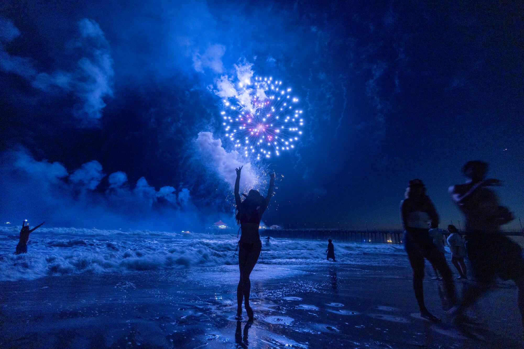 A woman, dances on the beach while watching the sky light up with blue fireworks.