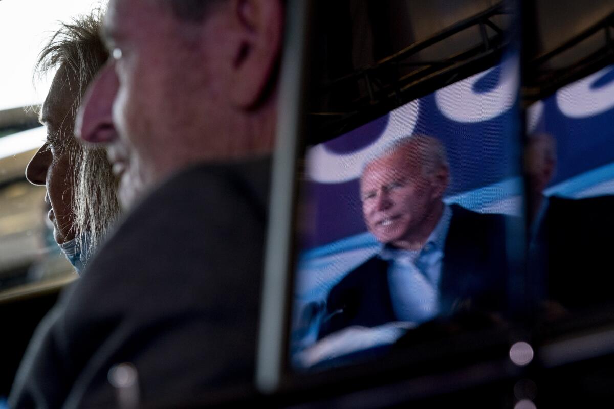 Joe Biden is seen reflected in a monitor at a rally where two supporters watch him from their car.