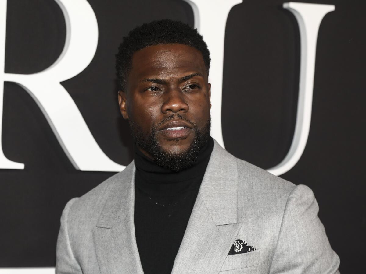 Kevin Hart wears a gray blazer and black turtleneck as he poses for photos at an event