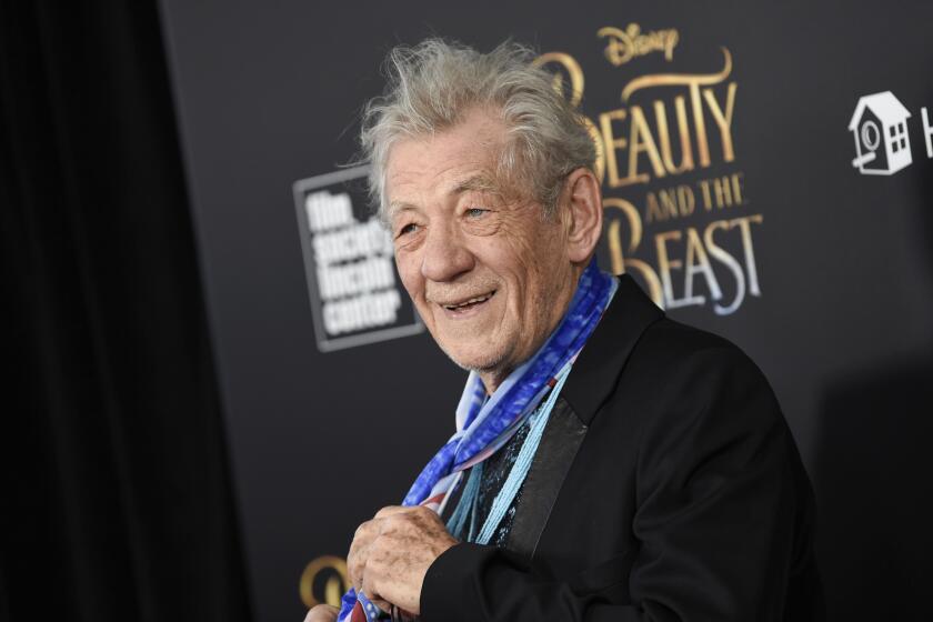 Ian McKellen in a black blazer, holding a blue neck scarf around his neck and posing against a black backdrop