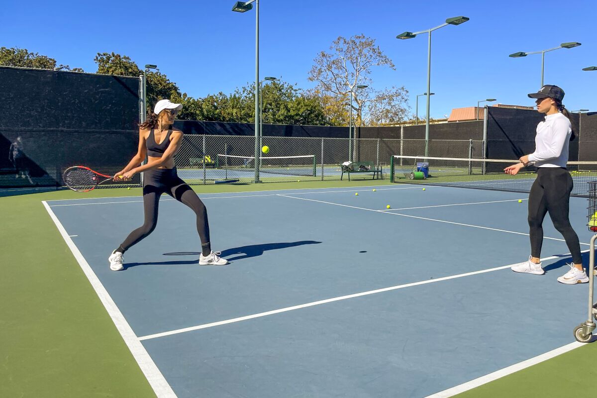 A woman plays tennis while another talks her through her swing