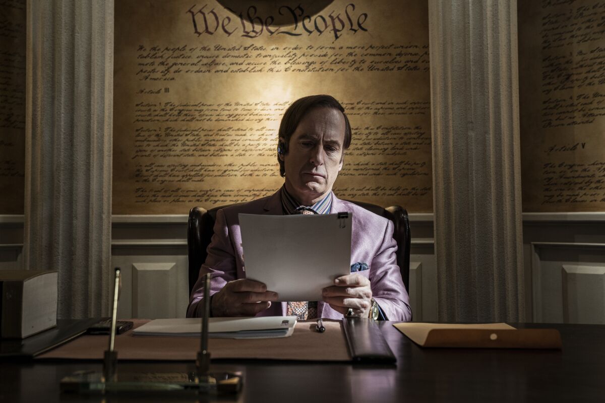 A lawyer in a pink suit sits at a wooden desk holding paperwork. An image of the Constitution fills the wall behind him.