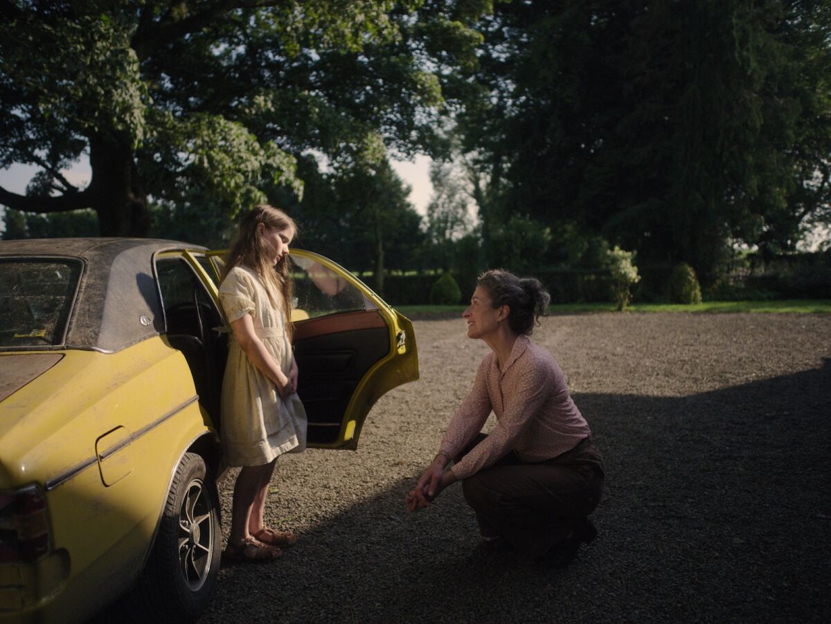 A girl stands near a car and is greeted by a woman who hunkers down to the girl's eye level.
