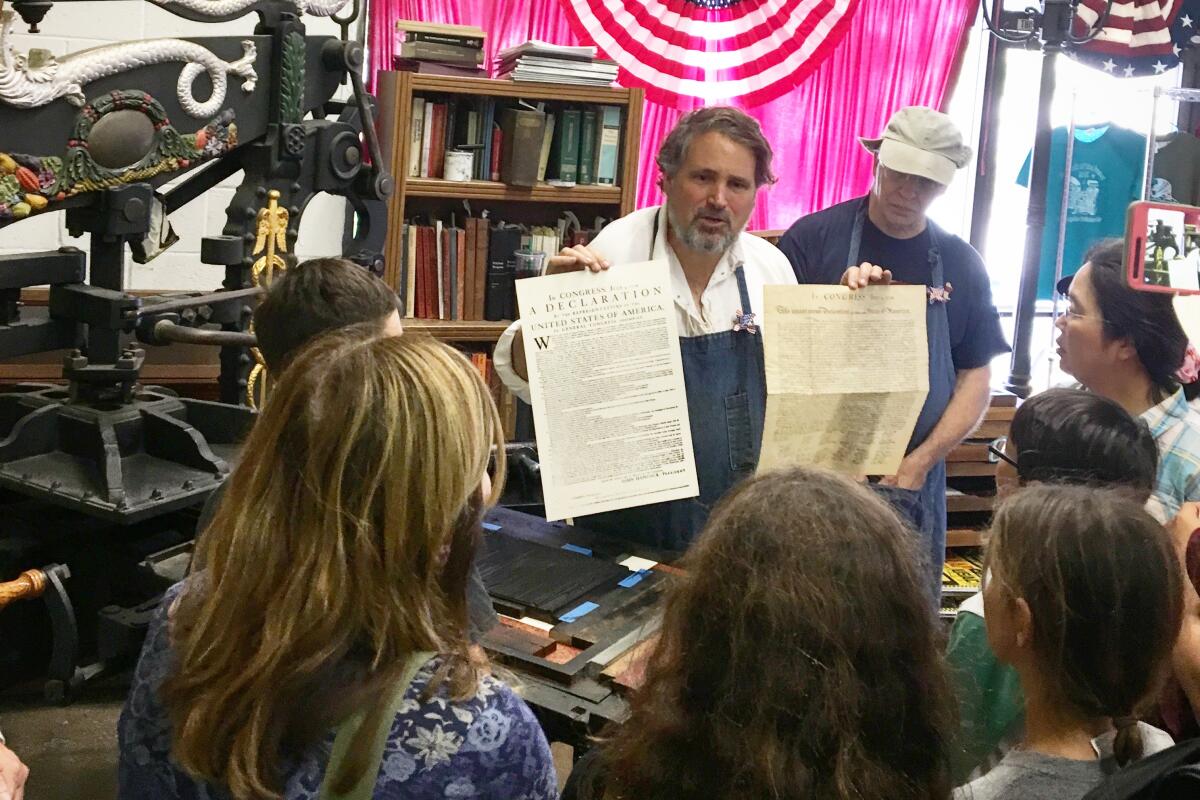 A man holds up two prints to a crowd of museum visitors during a printing press demonstration. 