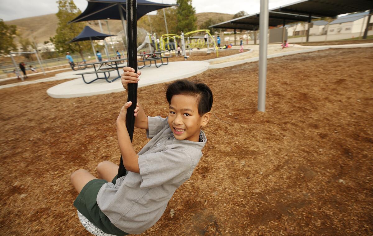 A smiling boy with dark hair, in a gray shirt and dark shorts, swings on a playground 