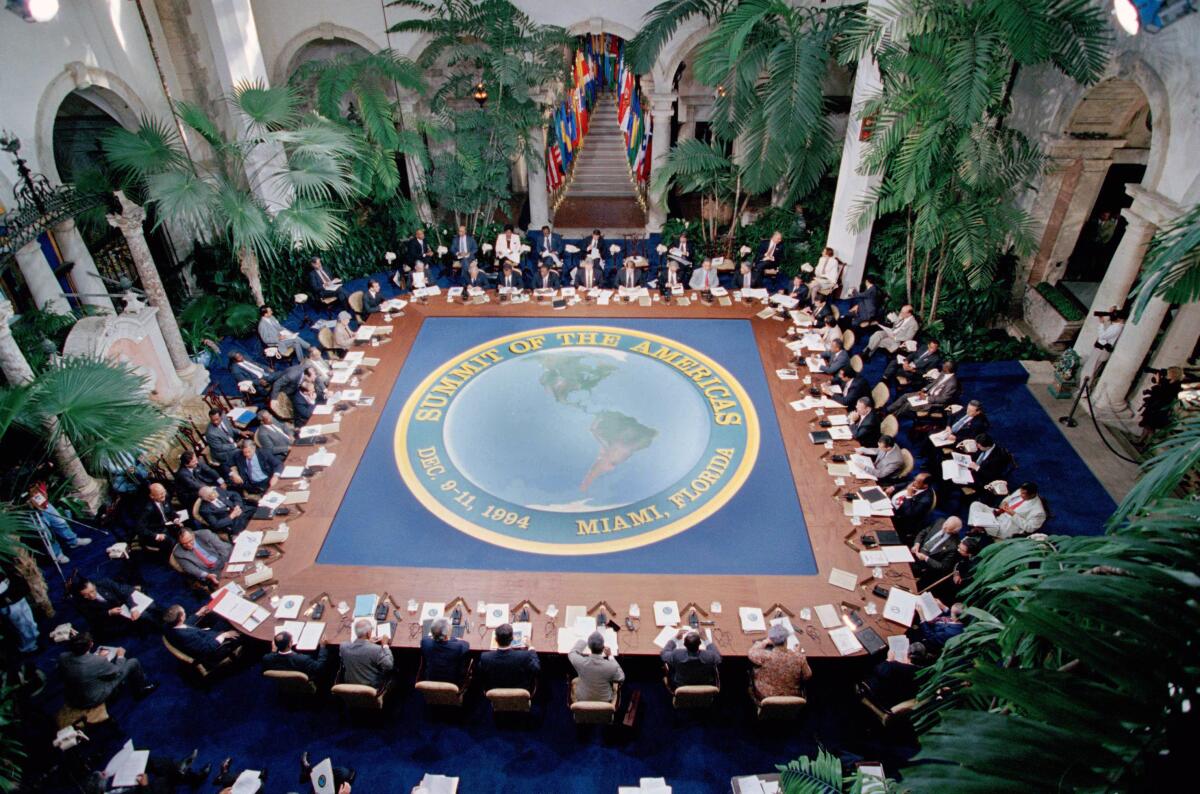 A view from above of people seated at a large table bearing a logo of a map and the words Summit of the Americas 