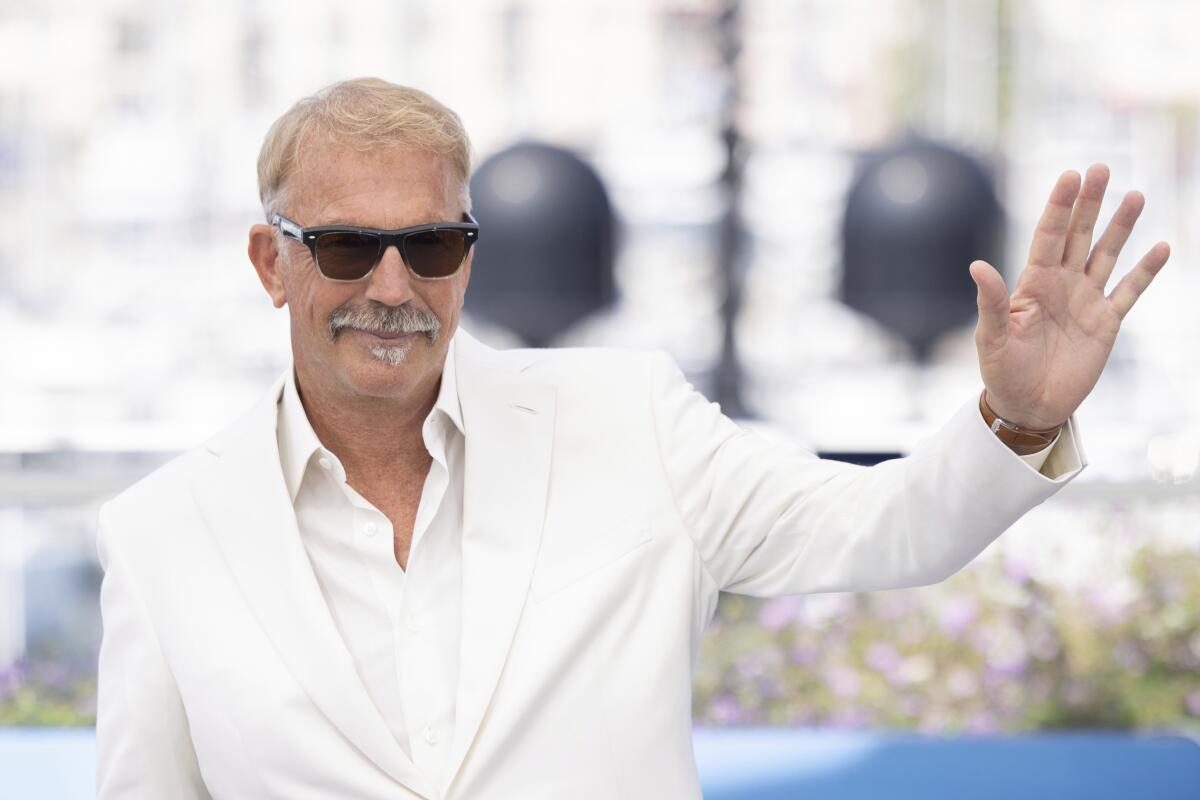 Kevin Costner waving in a white suit, white shirt and dark sunglasses