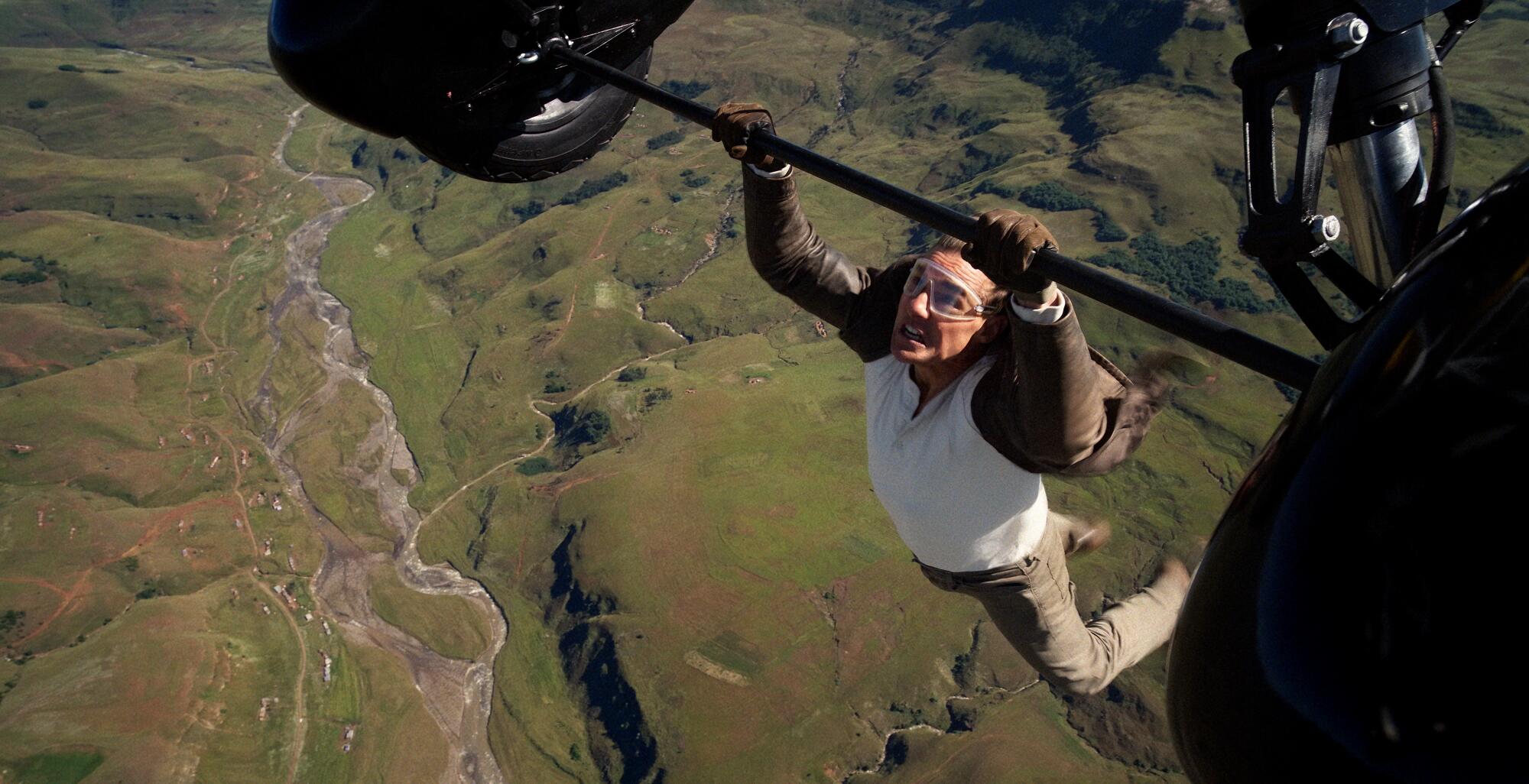 A man hangs from the wheels of a biplane high in the sky.