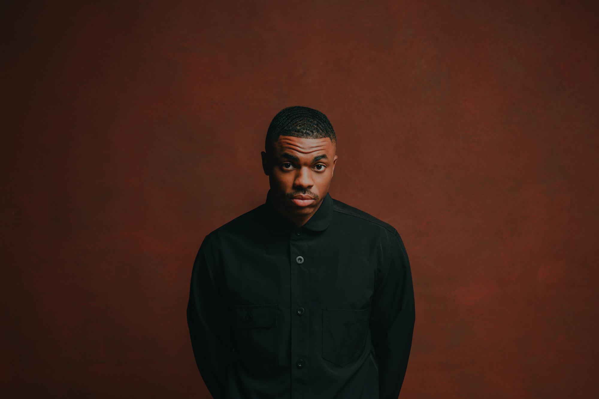 Vince Staples in black jacket and pants standing in front of a brown backdrop with his arms behind his back.