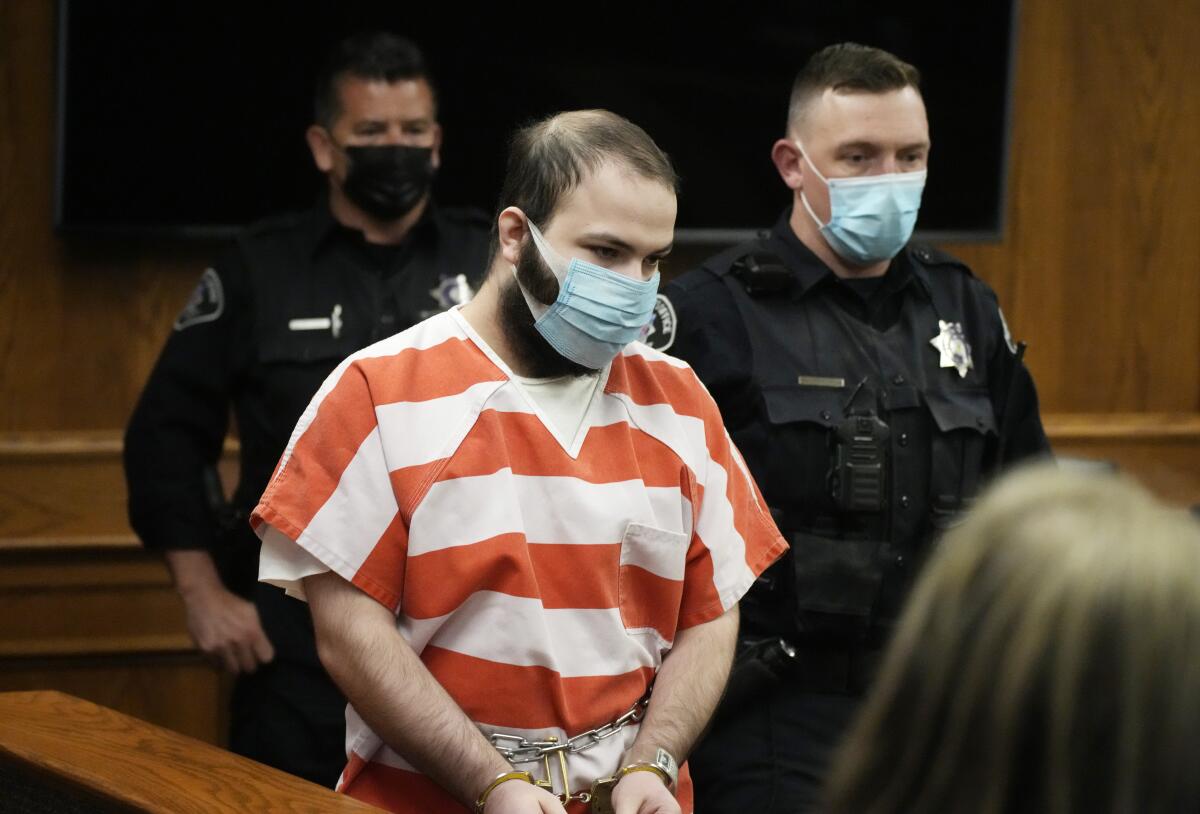 A man in handcuffs and red and white striped jail shirt is led into a courtroom.