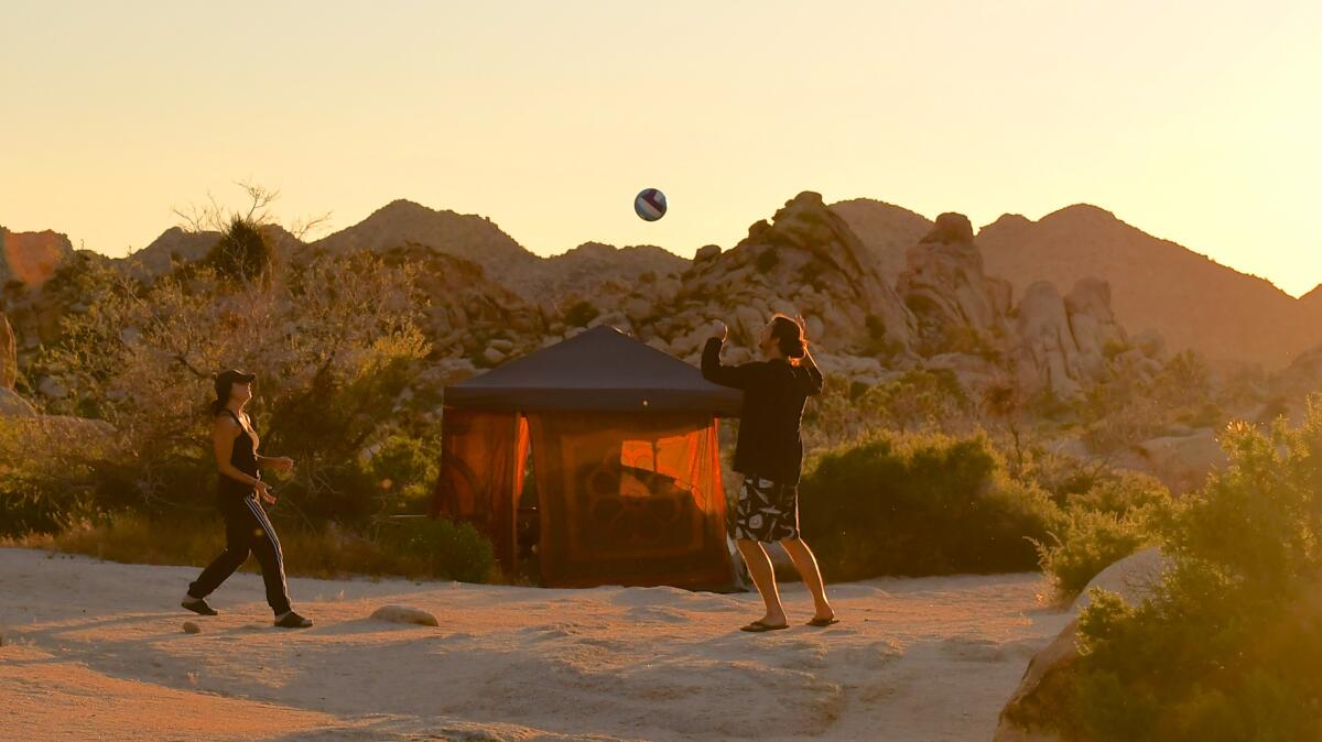 Two people play with a ball outside a tent backed by desert plants and mountain crags.