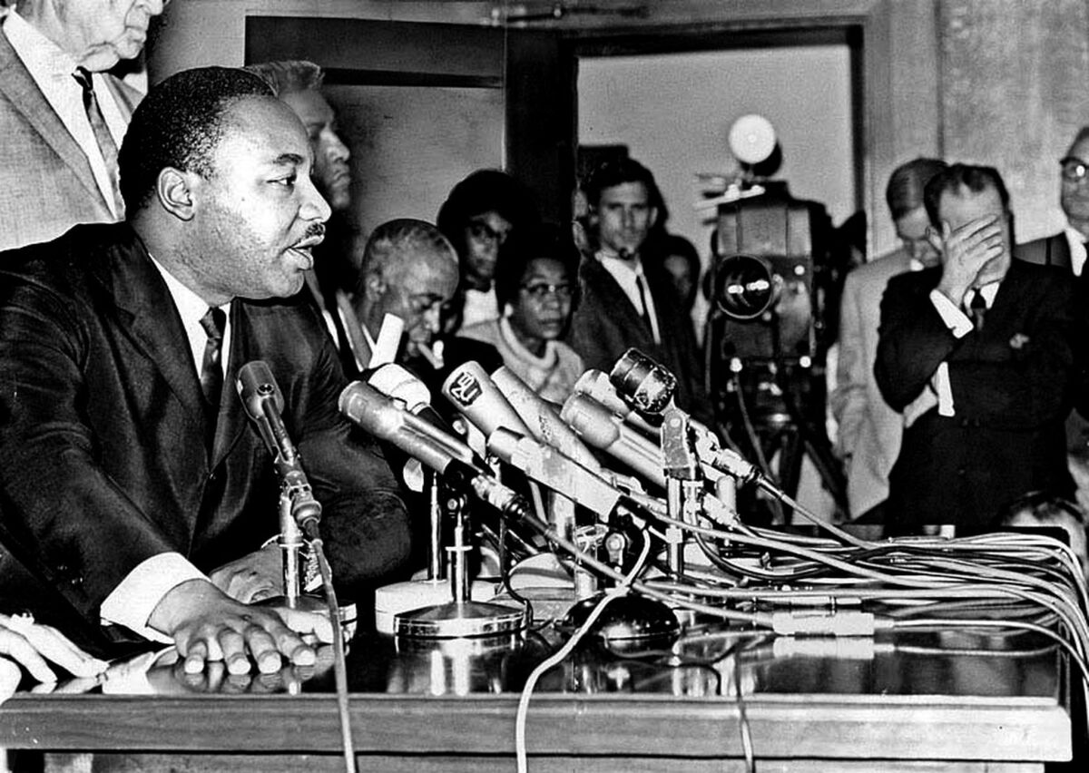 A black and white photo of a Black man speaking into a group of microphones in a crowded room