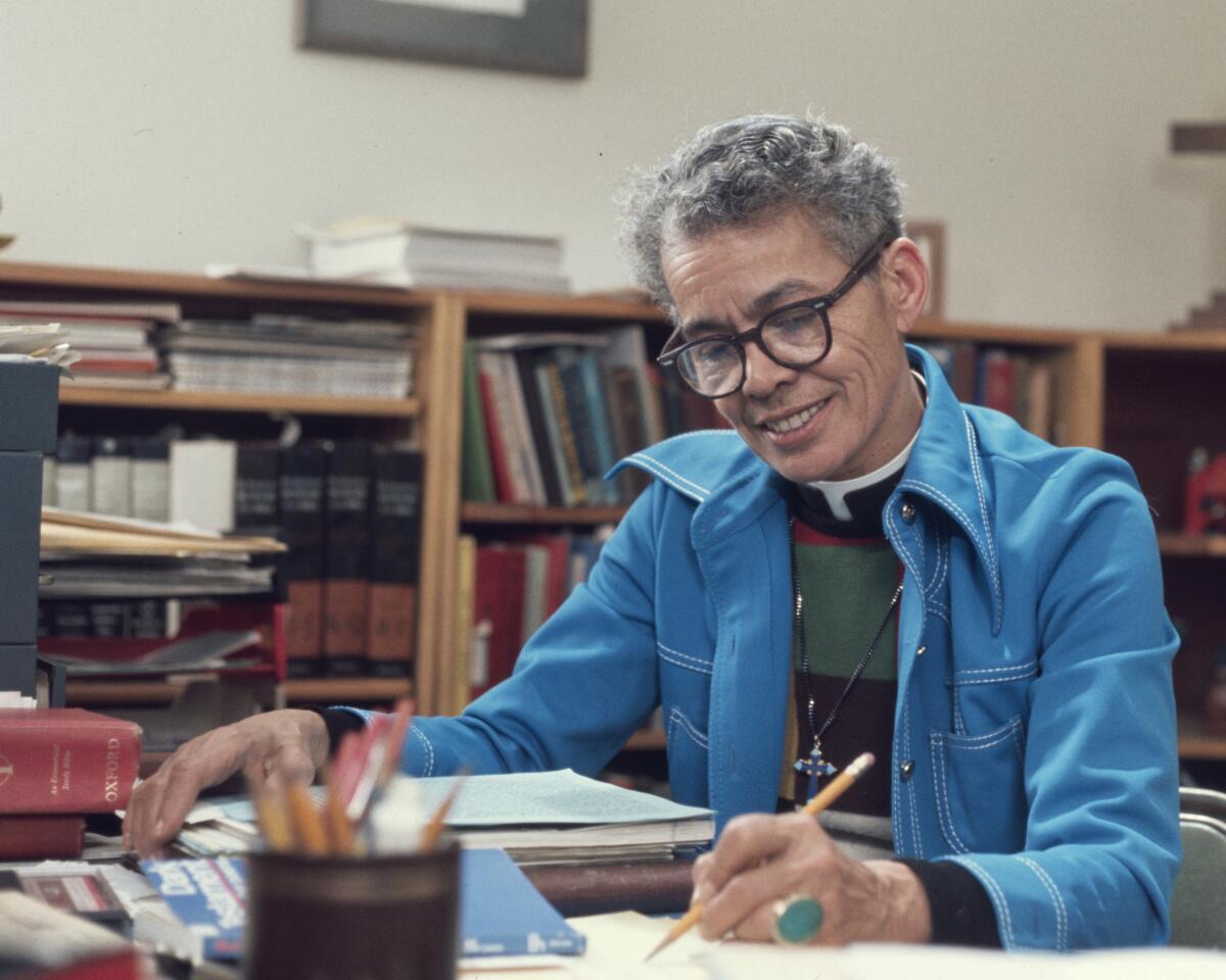 A person writes with a pencil at a table full of papers, with bookshelves behind.