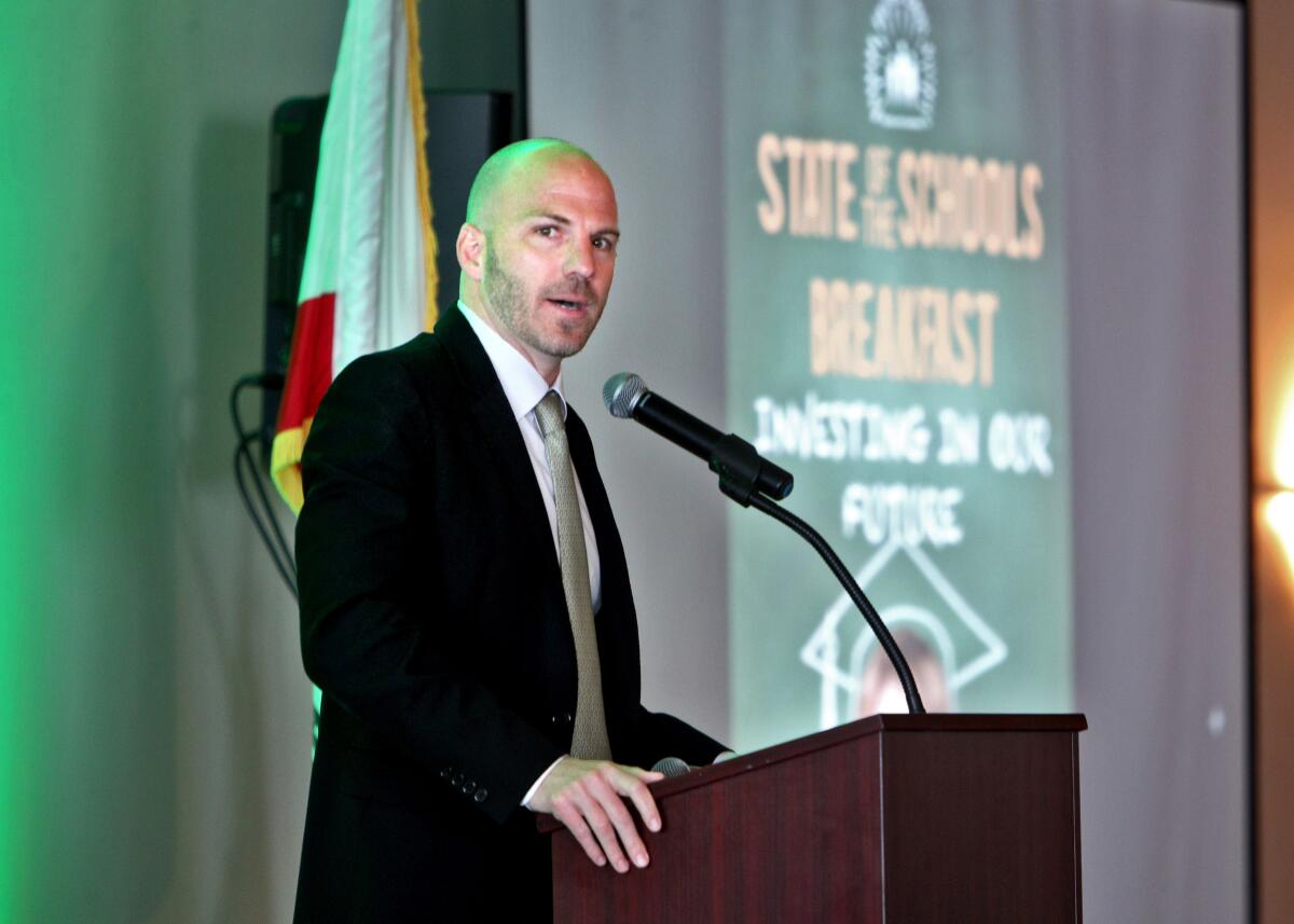 Framed waist up, a man in a suit stands on a podium and speaks from behind a lectern while looking toward the camera.