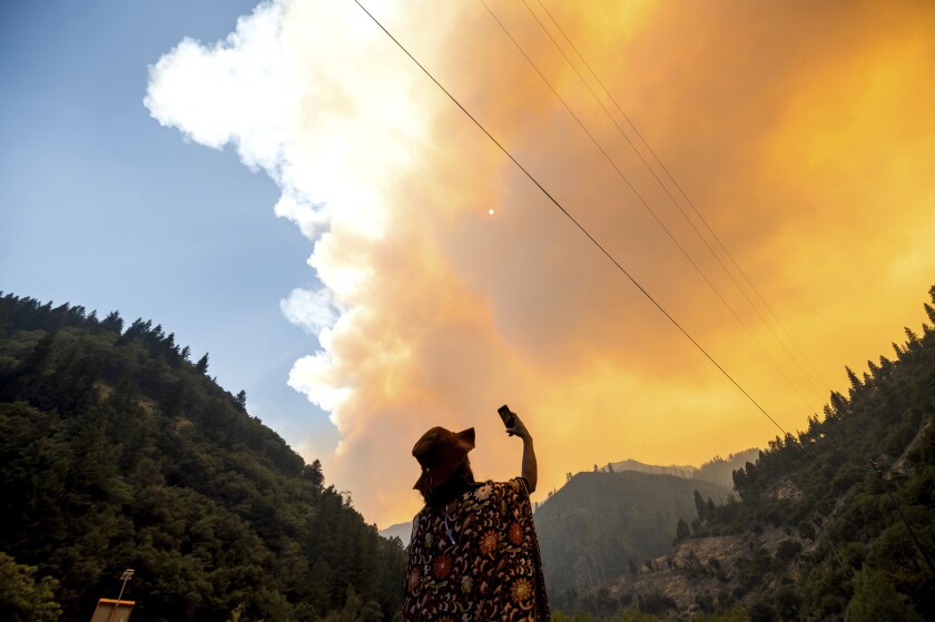 A woman takes a photo of smoke and flames as they rise over a mountain