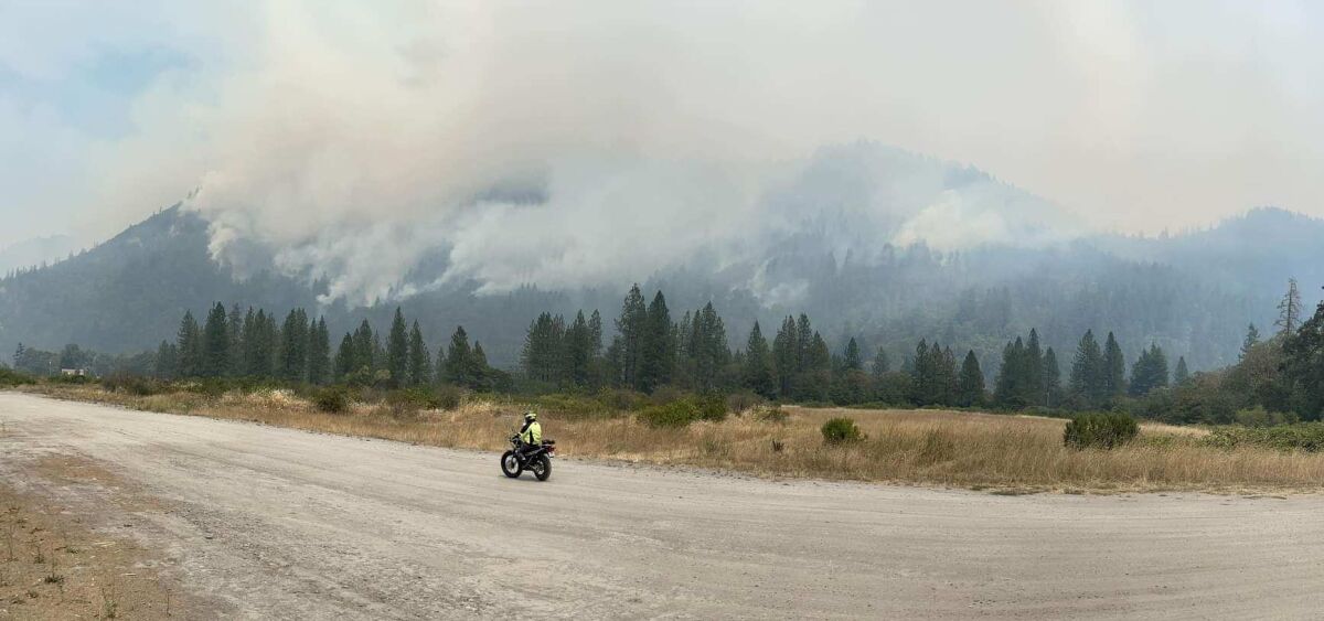 Smoke rises above trees and hills as a motorcyclist rides in the foreground.