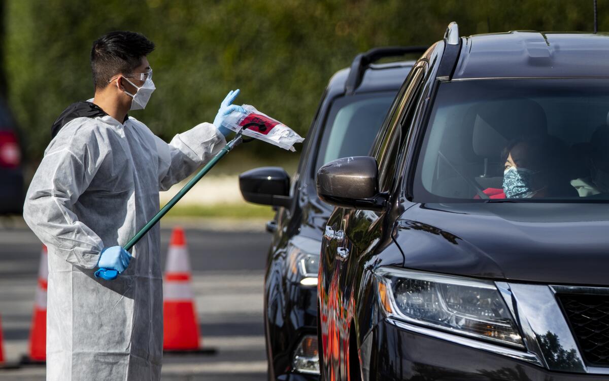 A man in a paper gown, gloves and mask holds a plastic package next to cars in a line.