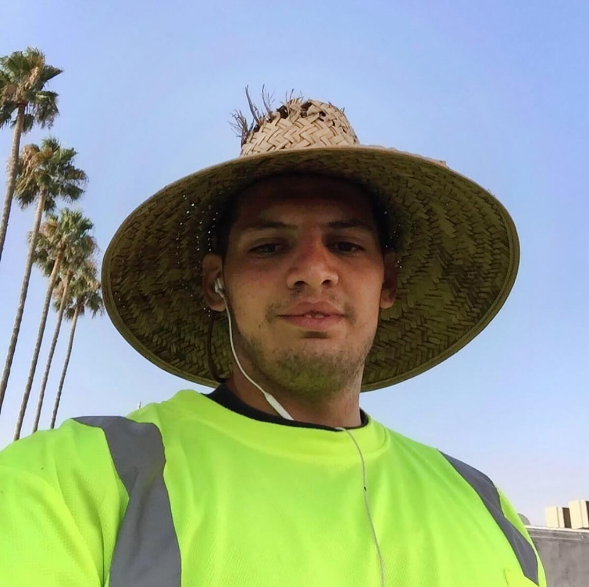 A self-portrait of Robert Brown wearing a neon yellow T-shirt, wired headphones, and a wicker hat with palm trees in the background.