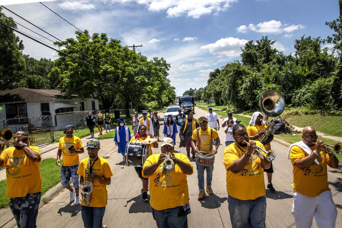 A marching band in yellow t-shirts marches down a residential street, followed by high school graduates in gowns