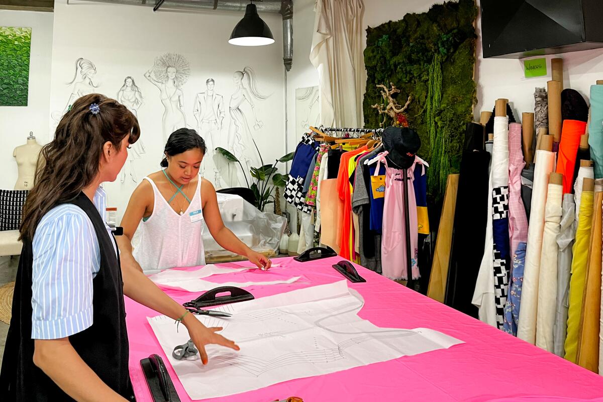 Two women look at sewing patterns on a bright pink table.