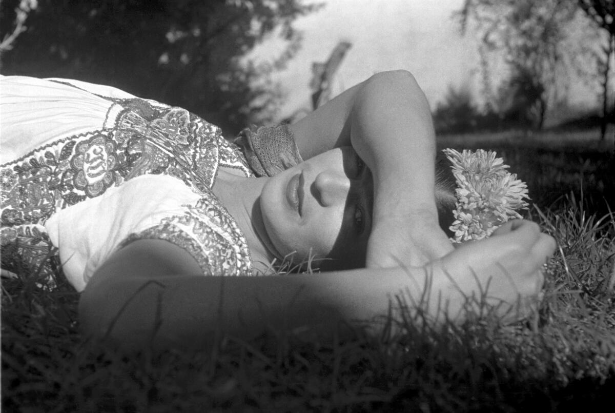 A black and white photo shows Frida Kahlo in a floral blouse, blocking the sun from her eyes while laying in the grass.