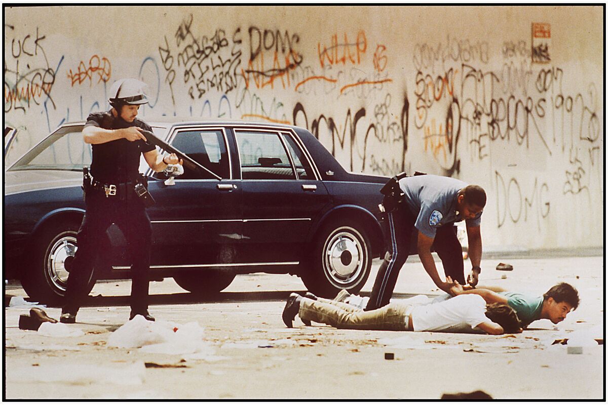 A police officer aims a rifle at two men lying on the ground.