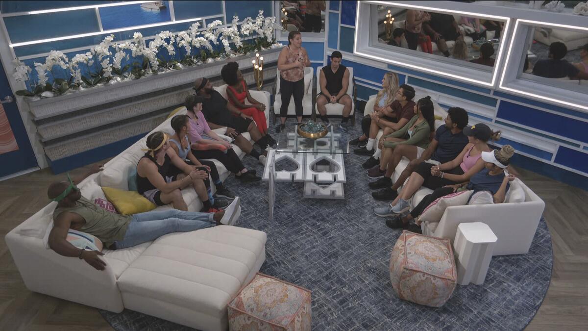 A large group of people sit around a living room set on the reality show "Big Brother"
