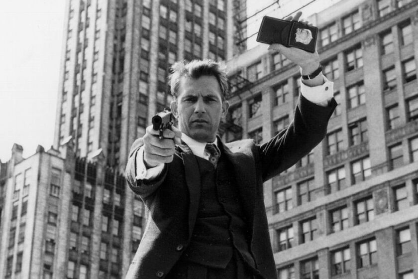 Kevin Costner holding a badge over his head and pointing a gun at the camera in "The Untouchables" (1987)