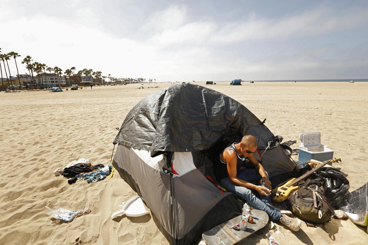 A homeless man sits near his belongings on a sandy beach.