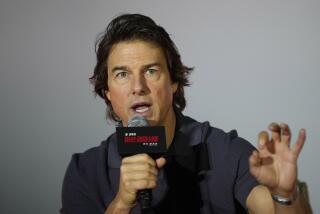 Tom Cruise is wearing a dark gray polo shirt and is sitting on a stage and holding a microphone while speaking