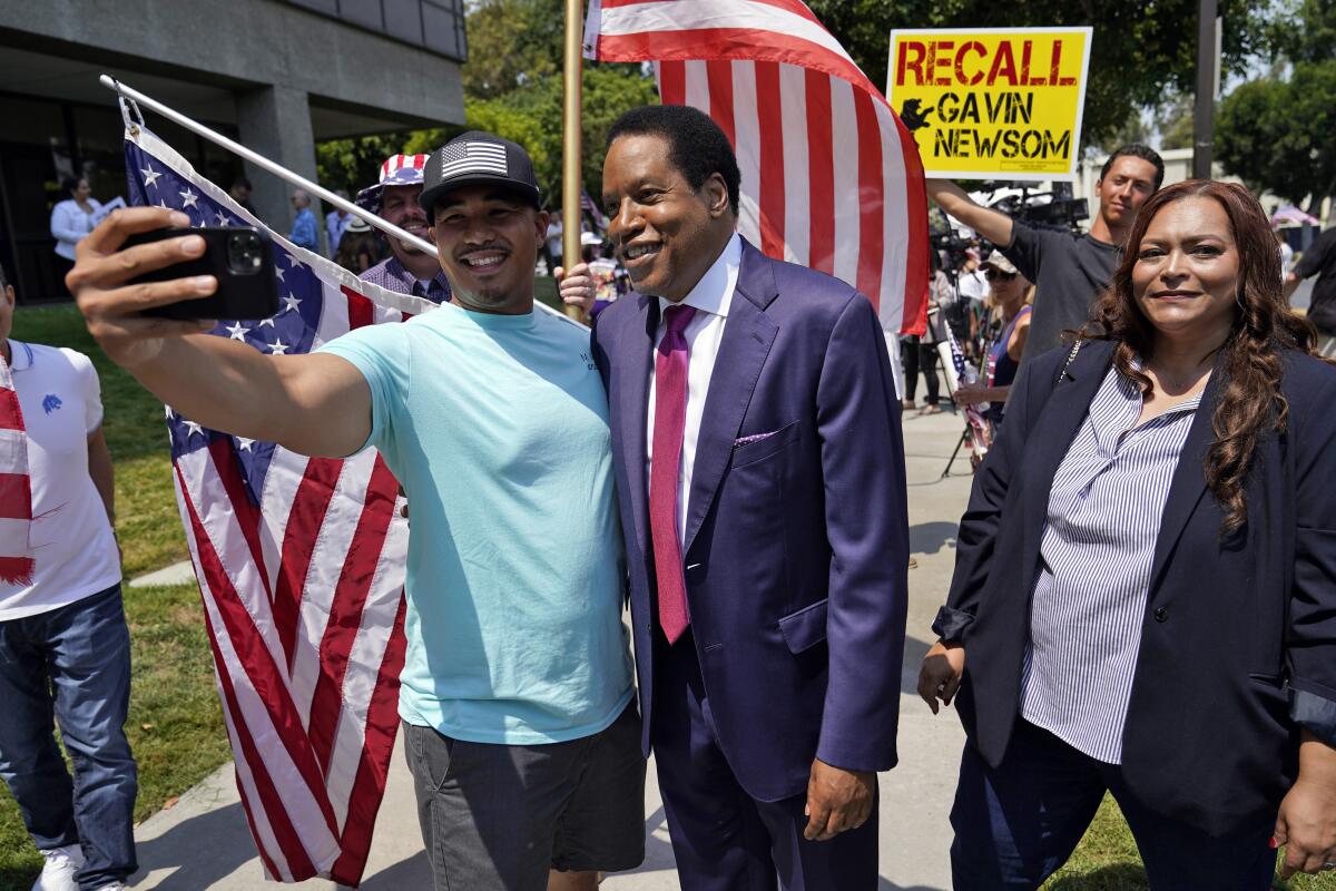 A man in a suit and red tie stands next to a man, left, taking their picture with a phone near a Recall Gavin Newsom sign.